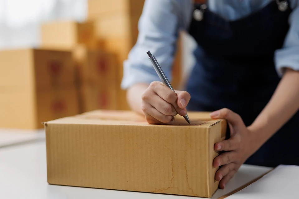 Entrep&ocirc;t logistique avec employ&eacute;e pr&eacute;parant commandes, utilisant pistolet d&eacute;vidoir rouge pour fermer colis carton marqu&eacute; "FRAGILE"